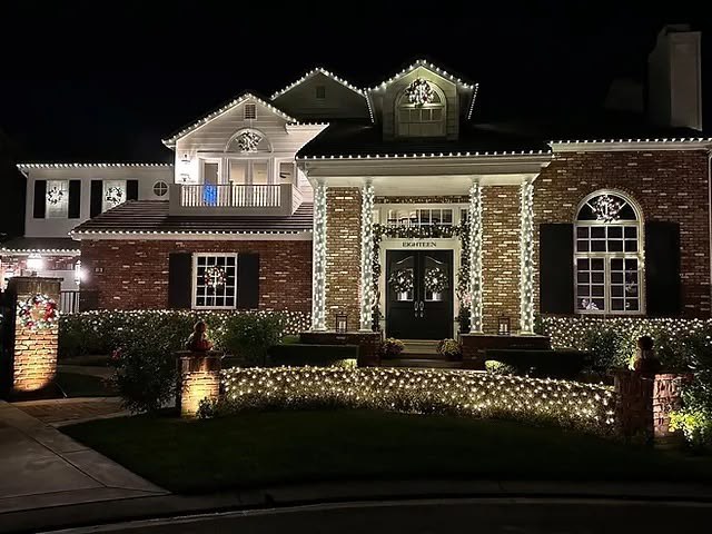 Brick home decorated with warm white roof lighting, illuminated columns, wreath and hedge lights.
