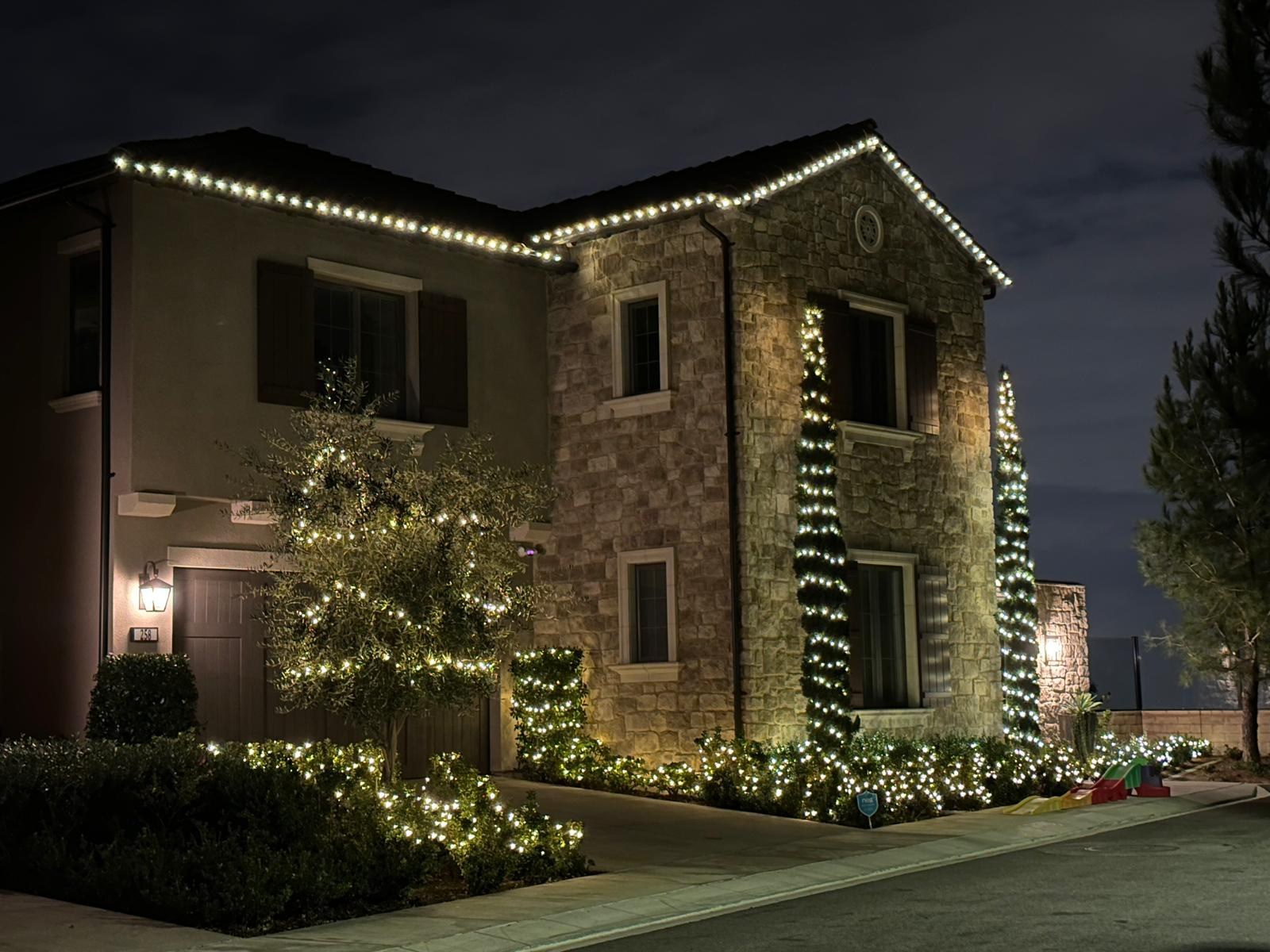 Completed warm white installation tracing a two-story home's roofline, wrapped columns and front garden.